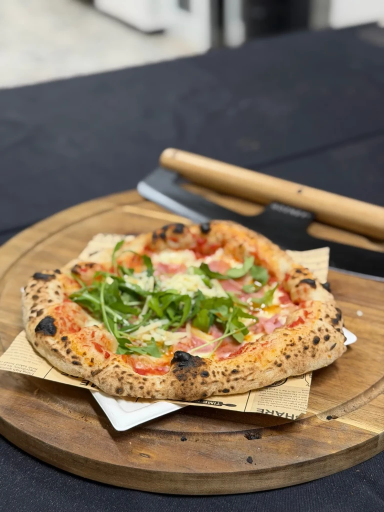Frisch gebackene neapolitanische Pizza mit Rucola und Schinken auf dem Holzbrett, daneben ein Pizzaschneider