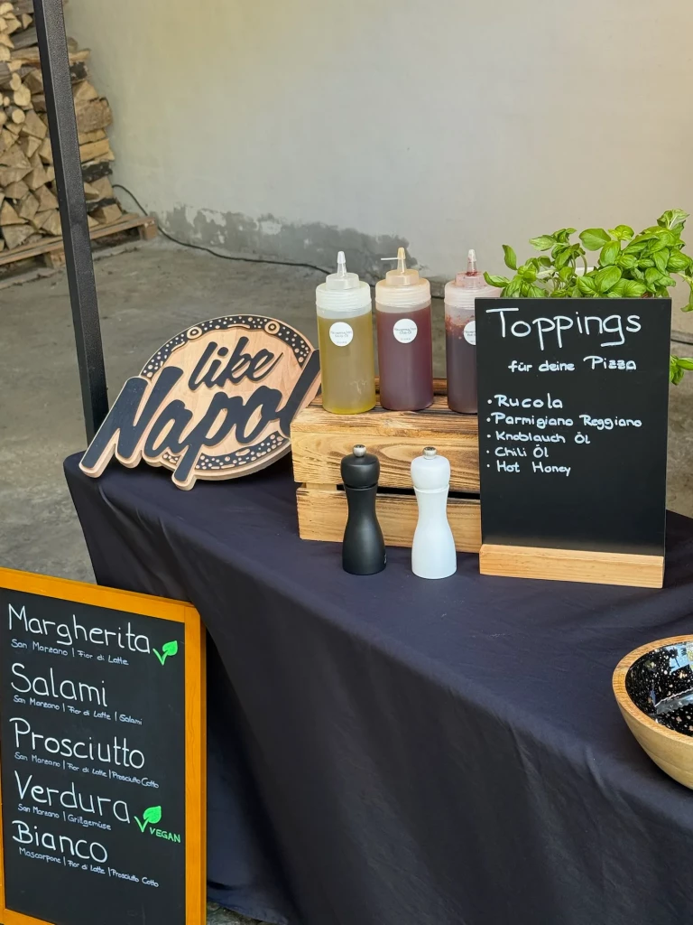 Toppings-Station mit Rucola, Parmigiano Reggiano und Ölen neben der Sortentafel und dem Like-Napoli-Logo