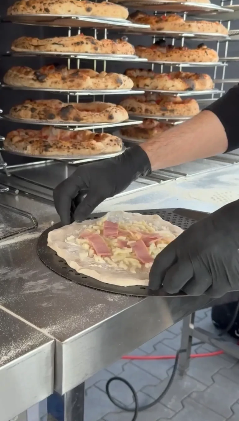 Pizzabäcker belegt eine Pizza, dahinter Etageren voller vorgebackener Pizzen