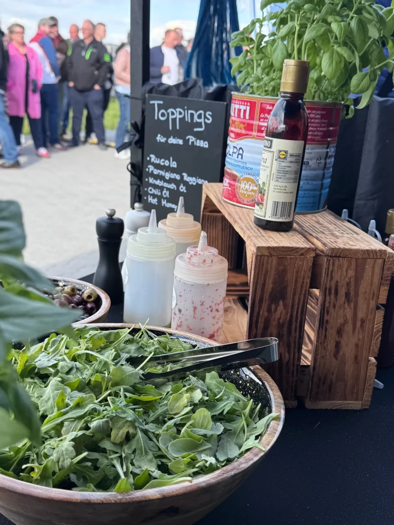 Toppings-Station mit frischem Rucola und Basilikum, im Hintergrund die Gäste des Polterabends